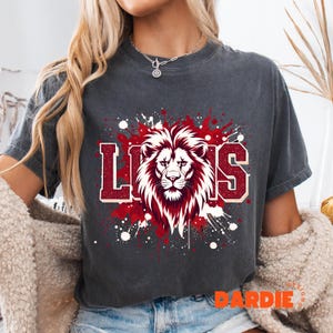 May include: Dark gray t-shirt featuring a lion's head graphic with the word "LIONS" in a bold, collegiate-style font. The design incorporates red and white accents with a splatter effect, creating a dynamic and eye-catching look. The shirt is a casual wear item.