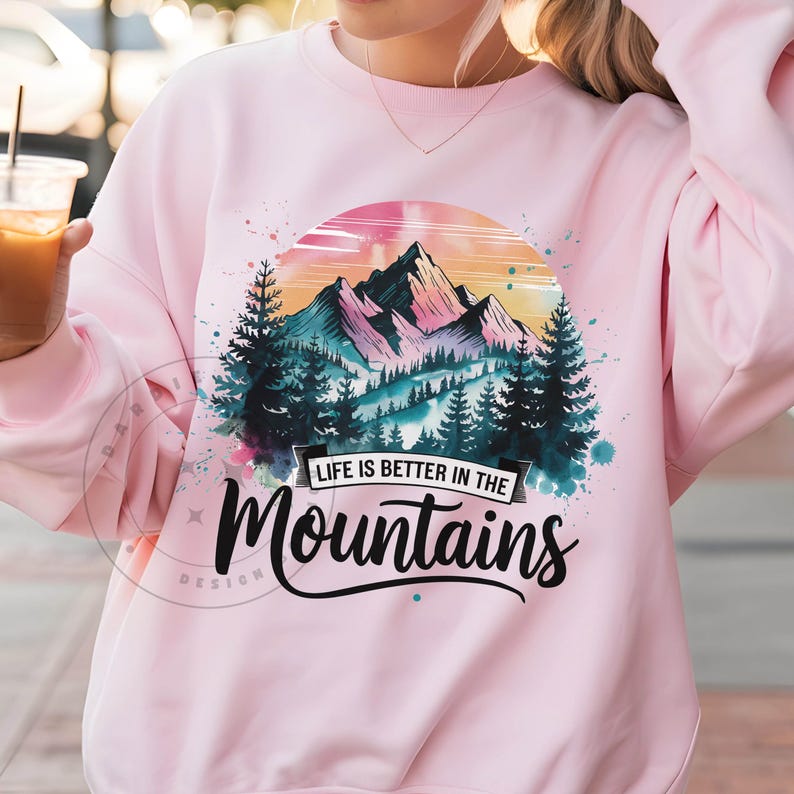 Op de afbeelding: Roze sweatshirt met een aquarelontwerp van bergen en bomen. De tekst "Life is better in the mountains" is op de voorkant gedrukt.