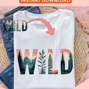 May include: White t-shirt with the word "WILD" in a nature-themed design. The letters feature illustrations of mountains, trees, and plants in shades of green, orange, and blue. The shirt is folded on top of blue jeans.