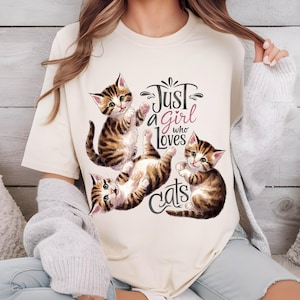 May include: A white t-shirt with the text "Just a girl who loves cats" printed on it. The text is in black with a pink heart. There are three cartoon cats in brown and white on the shirt.