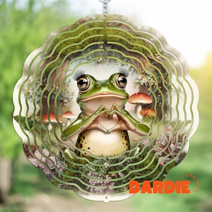 May include: A green frog with eyelashes and a heart-shaped hand gesture is featured on a metal wind spinner. The spinner has a layered design with a green, brown, and white background. The frog is surrounded by mushrooms and flowers.
