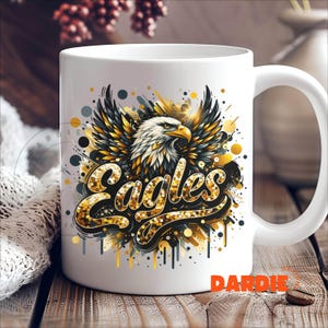 Peut inclure: Une tasse en c&eacute;ramique blanche avec un motif or et noir repr&eacute;sentant un aigle avec le mot "Eagles" en lettres dor&eacute;es paillet&eacute;es.