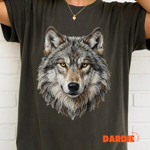 May include: Dark green t-shirt featuring a detailed illustration of a wolf's head. The wolf has gray, brown, and white fur with yellow eyes. The word "DARDIE" is printed in orange at the bottom of the shirt.