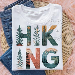 Op de afbeelding: Wit T-shirt met een grafisch ontwerp van het woord "HIKING" in kleurrijke letters. Elke letter is gevuld met een natuursc&egrave;ne, waaronder bomen, bergen en een meer.