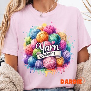 May include: A light pink t-shirt featuring a colorful graphic of yarn balls in various colors, including pink, blue, yellow, and green. The words "Yarn Addict" are displayed on a banner. The design has a watercolor effect.