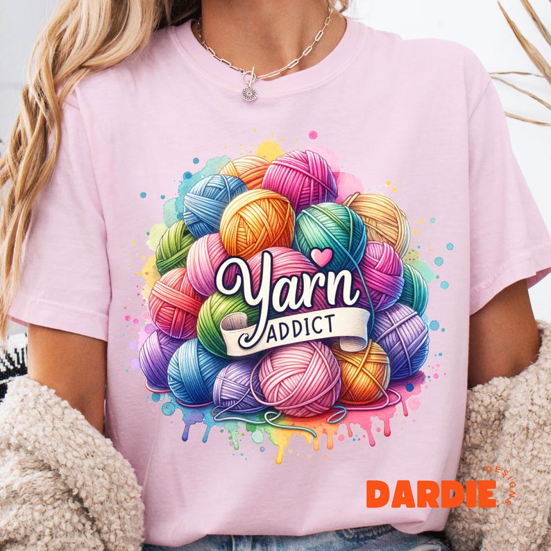 May include: A light pink t-shirt featuring a colorful graphic of yarn balls in various colors, including pink, blue, yellow, and green. The words "Yarn Addict" are displayed on a banner. The design has a watercolor effect.