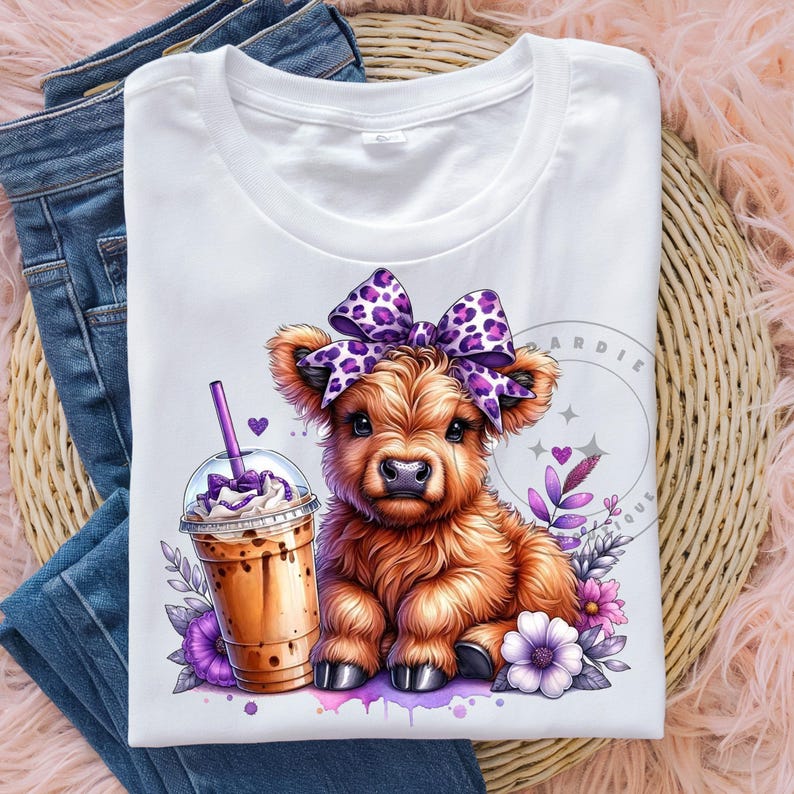 Pu&ograve; includere: Una maglietta bianca con un simpatico cartone animato di mucca Highland che indossa un fiocco viola e bianco a pois. La mucca tiene un drink al caff&egrave; freddo marrone e bianco. Lo sfondo &egrave; un design ad acquerello con fiori viola.