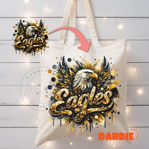 Peut inclure: Un sac fourre-tout en toile blanche avec un motif d'aigle dor&eacute; et noir. L'aigle est entour&eacute; de paillettes dor&eacute;es et le mot "Eagles" est &eacute;crit en lettres dor&eacute;es paillet&eacute;es. Le sac a des anses noires.