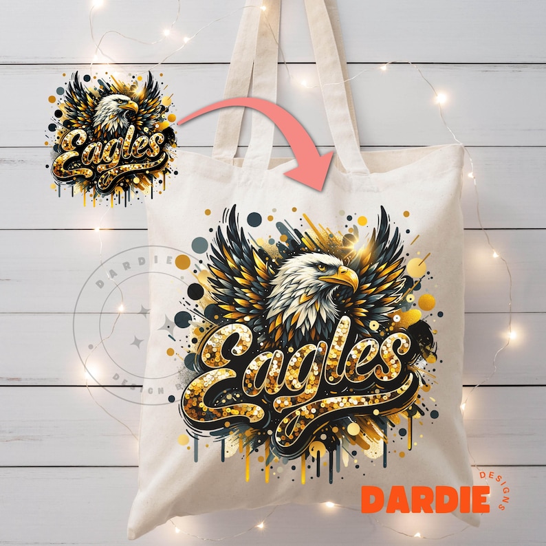 Peut inclure: Un sac fourre-tout en toile blanche avec un motif d'aigle dor&eacute; et noir. L'aigle est entour&eacute; de paillettes dor&eacute;es et le mot "Eagles" est &eacute;crit en lettres dor&eacute;es paillet&eacute;es. Le sac a des anses noires.