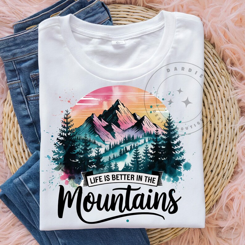 Op de afbeelding: Wit T-shirt met een aquarelontwerp van bergen en bomen. De tekst "Life is better in the mountains" is op het T-shirt gedrukt.