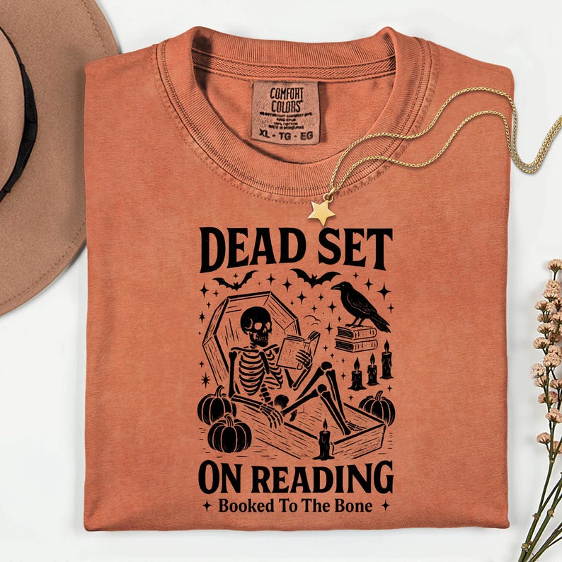 May include: An orange t-shirt with the text "DEAD SET ON READING + Booked To The Bone +" and a graphic of a skeleton reading in a coffin, surrounded by pumpkins, bats, and candles. A gold necklace with a star pendant is draped across the shirt.