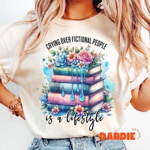 Puede incluir: Camiseta color crema con un gráfico de una pila de libros con detalles florales y el texto "Crying Over Fictional People is a Lifestyle." El diseño incluye colores azul, rosa y verde.