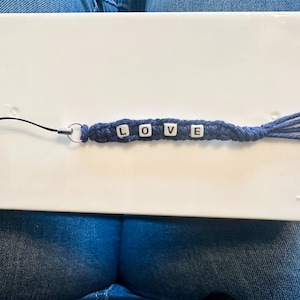 May include: A blue macrame wrist strap with the word "LOVE" spelled out in white letter beads. The strap has a black cord loop on one end and a tassel on the other. The strap is lying on a white surface.