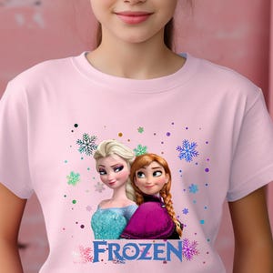 Puede incluir: Camiseta rosa con un gr&aacute;fico de los personajes Elsa y Anna de la pel&iacute;cula Frozen. El texto "FROZEN" est&aacute; impreso debajo de la imagen.