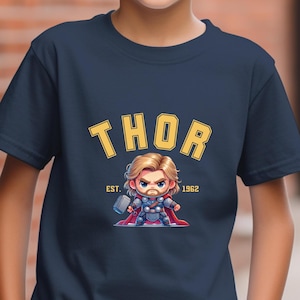 May include: Navy blue t-shirt featuring a cartoon illustration of Thor holding a hammer. The word "THOR" is printed in yellow arched lettering above the image, with "EST. 1962" below. The shirt is worn by a person wearing a black baseball cap.