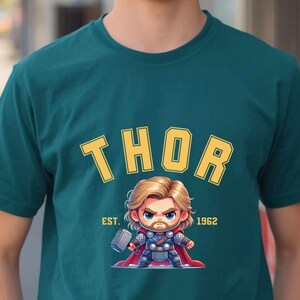 K&ouml;nnte beinhalten: T&uuml;rkisfarbenes T-Shirt mit gelbem, bogenf&ouml;rmigem "THOR"-Schriftzug und einer Cartoon-Illustration von Thor mit einem Hammer. Der Text "EST. 1962" steht darunter. Das T-Shirt wird von einer Person getragen.