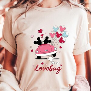 May include: Beige t-shirt featuring a pink car with two silhouettes and heart-shaped balloons. The text "Lovebug" is written in red script below the car. The design is a Valentine's Day theme.