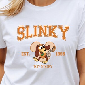 May include: White t-shirt featuring the word "SLINKY" in orange block letters above a cartoon image of Slinky Dog from Toy Story. The shirt also includes "EST. 1995" and "TOY STORY" in orange text.