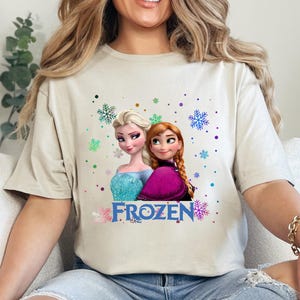 May include: A white t-shirt with a graphic of the characters Elsa and Anna from the movie Frozen. The graphic includes the word "FROZEN" and snowflakes.
