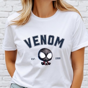 May include: White t-shirt featuring the word "VENOM" in navy blue arched lettering above a cartoon Venom character. The character is black with white eyes and a red mouth. The text "EST. 1988" is below the character.