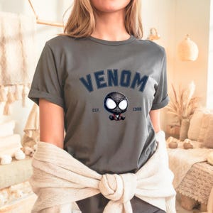 May include: Gray t-shirt with the word "VENOM" in navy blue arched text above a cartoon character with a black and white spider-like design. The text "EST. 1988" is below the character.