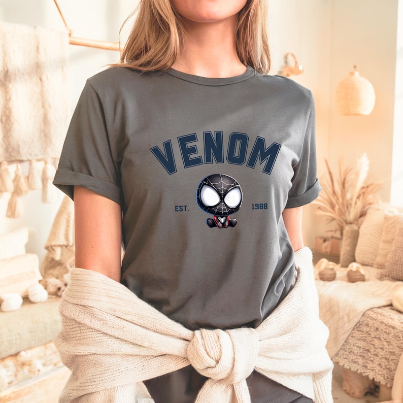 May include: Gray t-shirt with the word "VENOM" in navy blue arched text above a cartoon character with a black and white spider-like design. The text "EST. 1988" is below the character.