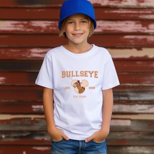 May include: White t-shirt featuring the word "BULLSEYE" in orange lettering, with an image of a cartoon horse and the text "EST. 1999 TOY STORY." The shirt is worn by a person wearing a blue cap.