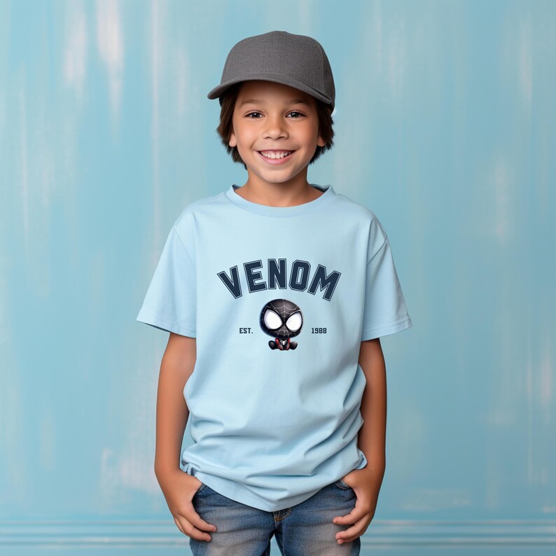 May include: Light blue t-shirt with the word "VENOM" in navy blue arched text. Below the text is a cartoon Venom character and the text "EST. 1988". The shirt is worn by a child wearing a gray baseball cap.