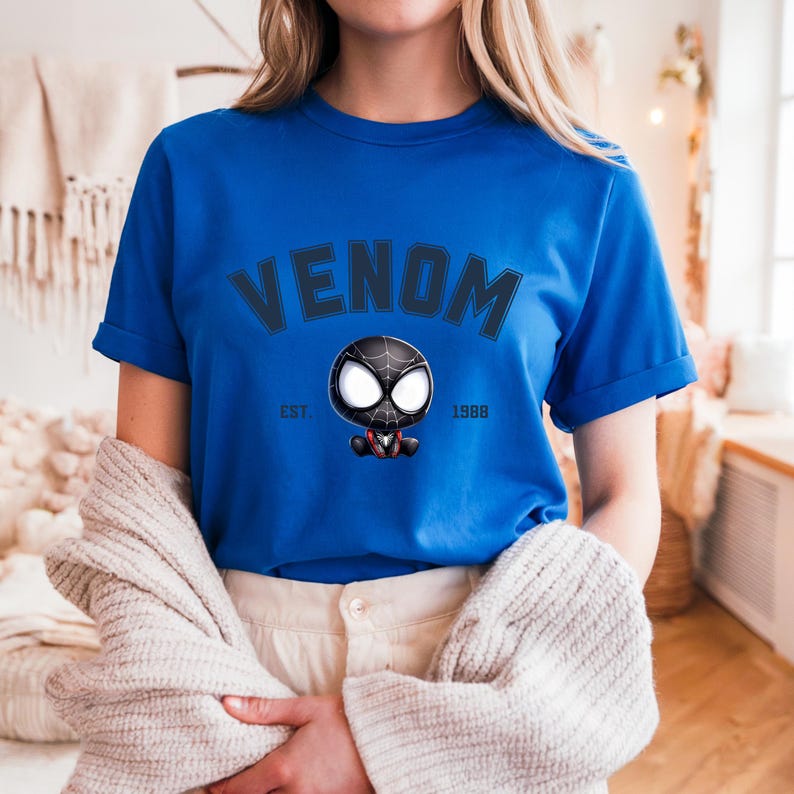 May include: Royal blue t-shirt featuring the word "VENOM" in a dark blue arched font. Below the word "VENOM" is a cartoon image of Venom. The text "EST. 1988" is printed below the image. The shirt has short sleeves.