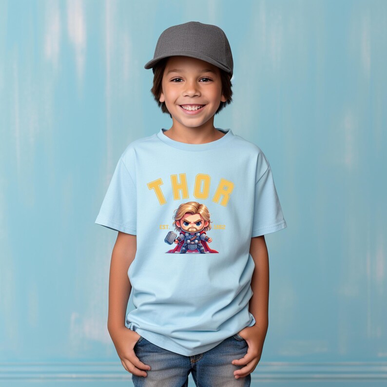 K&ouml;nnte beinhalten: Hellblaues T-Shirt mit dem Wort "THOR" in gelben Blockbuchstaben. Darunter eine Cartoon-Illustration von Thor, der einen Hammer h&auml;lt. Das T-Shirt wird von einem Kind mit einer grauen Baseballkappe getragen.