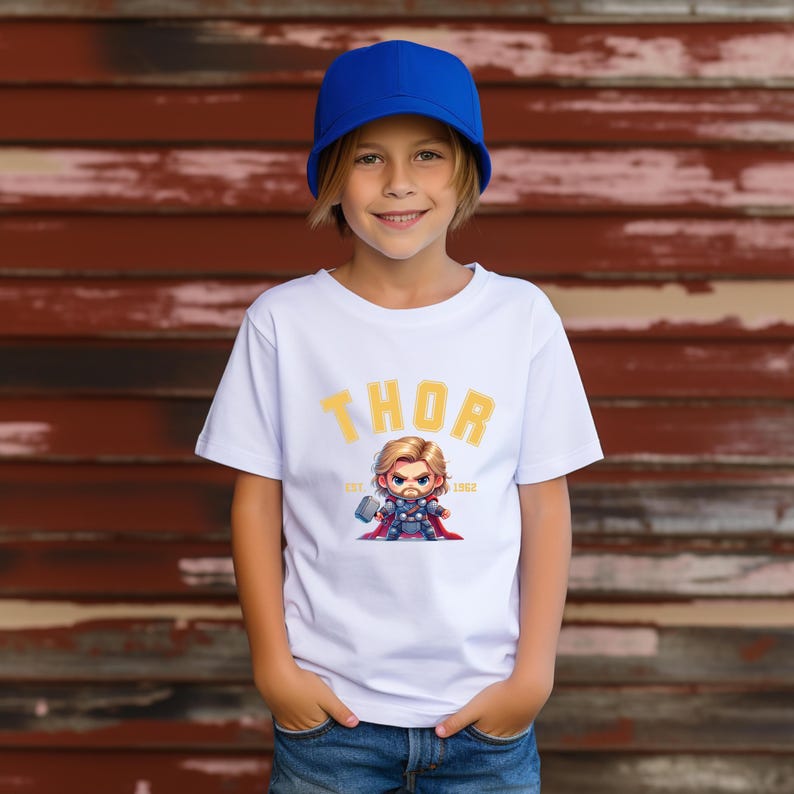 K&ouml;nnte beinhalten: Wei&szlig;es T-Shirt mit einer Cartoon-Illustration von Thor und dem Wort "THOR" in Gelb. Das Shirt hat auch den Text "EST. 1962". Die Person im Bild tr&auml;gt eine blaue Baseballkappe.