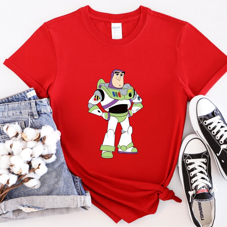 May include: A red t-shirt featuring a cartoon image of Buzz Lightyear from Toy Story. The character is depicted in his signature green, white, and purple spacesuit. The shirt is paired with denim shorts and black Converse sneakers.