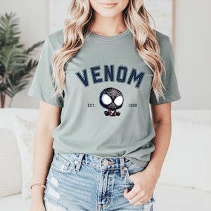 May include: Sage green t-shirt featuring the word "VENOM" in navy blue arched lettering above a cartoon Venom character. The graphic includes the text "EST. 1988" on either side of the character. The shirt is made of a soft material.