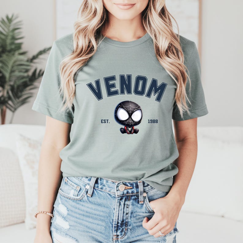 May include: Sage green t-shirt featuring the word "VENOM" in navy blue arched lettering above a cartoon Venom character. The graphic includes the text "EST. 1988" on either side of the character. The shirt is made of a soft material.