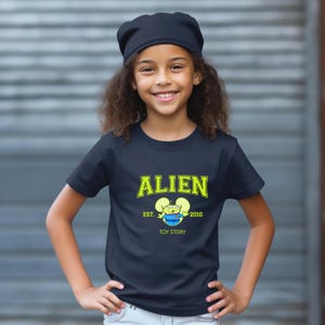 May include: A navy blue t-shirt with the word "ALIEN" in yellow block letters. Below the word "ALIEN" is a cartoon alien character and the text "EST. 2010 TOY STORY". The shirt is worn by a young person.
