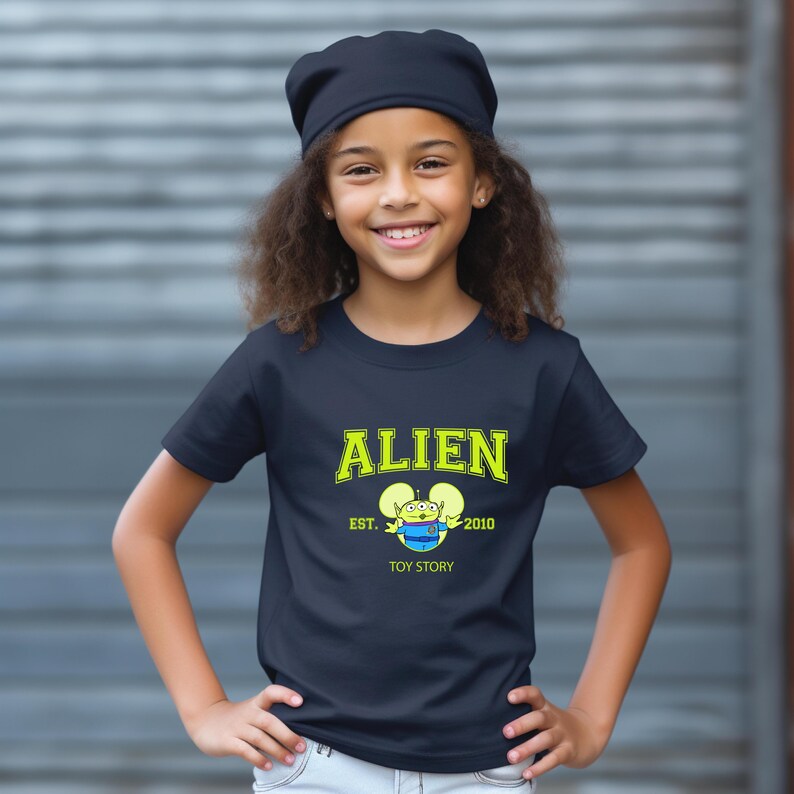 May include: A navy blue t-shirt with the word "ALIEN" in yellow block letters. Below the word "ALIEN" is a cartoon alien character and the text "EST. 2010 TOY STORY". The shirt is worn by a young person.