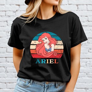 Puede incluir: Camiseta negra con un gr&aacute;fico de estilo vintage de Ariel de La Sirenita. El gr&aacute;fico presenta a Ariel con cabello rojo, un sujetador de concha azul y un fondo de puesta de sol con el texto "ARIEL".