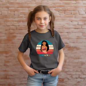 Puede incluir: Una camiseta gris oscuro con un gr&aacute;fico de estilo vintage de Moana, una princesa polinesia, con un fondo de puesta de sol. El texto "MOANA" est&aacute; impreso debajo del gr&aacute;fico.