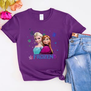 Puede incluir: Camiseta morada con un gr&aacute;fico de Elsa y Anna de la pel&iacute;cula Frozen. El gr&aacute;fico incluye la palabra "Frozen" en una fuente colorida.