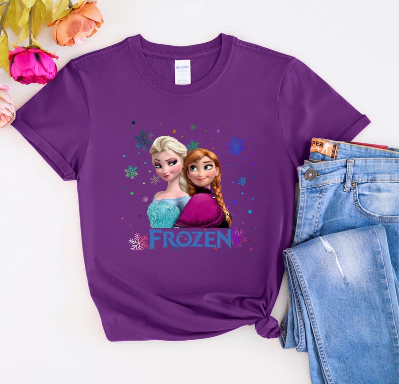 Puede incluir: Camiseta morada con un gr&aacute;fico de Elsa y Anna de la pel&iacute;cula Frozen. El gr&aacute;fico incluye la palabra "Frozen" en una fuente colorida.