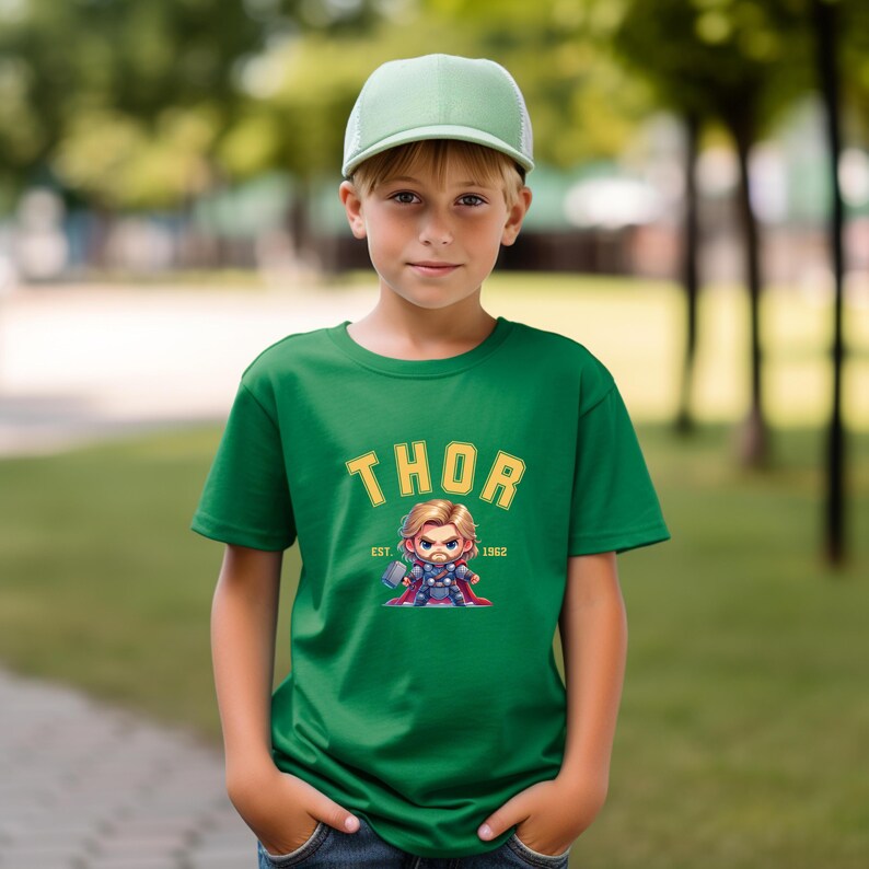 K&ouml;nnte beinhalten: Ein gr&uuml;nes T-Shirt mit dem Wort "THOR" in goldener Schrift und einer Cartoon-Illustration von Thor. Das Shirt hat auch "EST. 1962". Die Person auf dem Bild tr&auml;gt eine gr&uuml;ne Baseballkappe. Der Hintergrund ist unscharf.