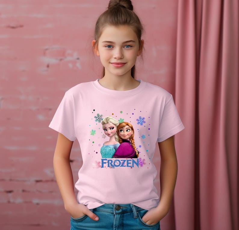 Puede incluir: Camiseta rosa con un gr&aacute;fico de los personajes Elsa y Anna de la pel&iacute;cula Frozen. El texto "FROZEN" est&aacute; impreso debajo de la imagen.