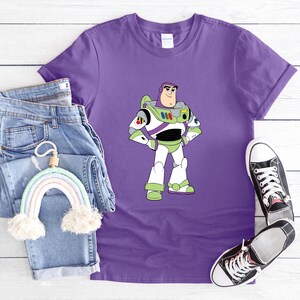 May include: Purple t-shirt featuring a cartoon image of Buzz Lightyear in his space suit. The shirt is paired with blue jeans, a rainbow-shaped wall hanging, and black canvas sneakers. The shirt is made of a soft material.