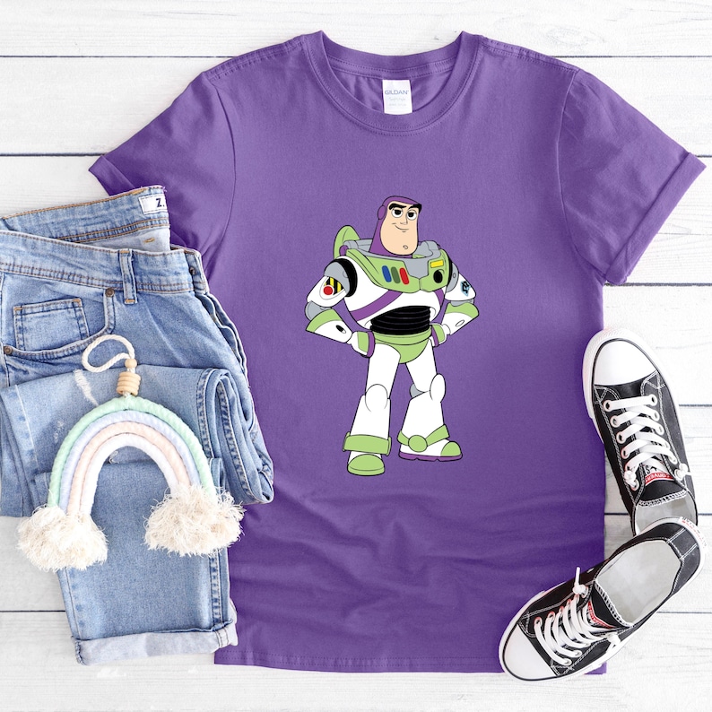 May include: Purple t-shirt featuring a cartoon image of Buzz Lightyear in his space suit. The shirt is paired with blue jeans, a rainbow-shaped wall hanging, and black canvas sneakers. The shirt is made of a soft material.
