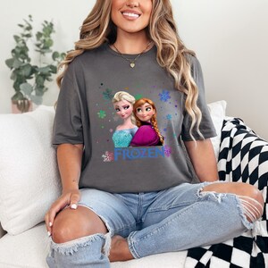 Puede incluir: Una camiseta gris con un gr&aacute;fico de los personajes Elsa y Anna de la pel&iacute;cula Frozen. El gr&aacute;fico incluye la palabra "FROZEN" y copos de nieve.