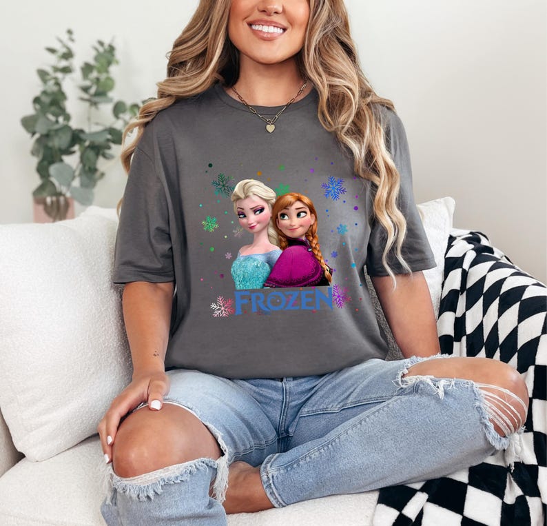 Puede incluir: Una camiseta gris con un gr&aacute;fico de los personajes Elsa y Anna de la pel&iacute;cula Frozen. El gr&aacute;fico incluye la palabra "FROZEN" y copos de nieve.