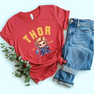 K&ouml;nnte beinhalten: Ein rotes T-Shirt mit dem Wort "THOR" in gelben Blockbuchstaben und einer Cartoon-Illustration von Thor. Das Shirt wird mit hellblauen Jeans kombiniert. Der Text "EST. 1962" ist ebenfalls sichtbar.