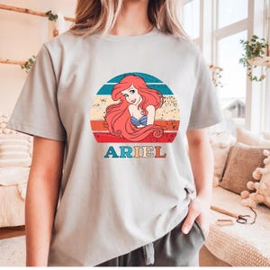 Puede incluir: Una camiseta gris claro con un gr&aacute;fico de estilo vintage de Ariel de La Sirenita. El gr&aacute;fico presenta a Ariel con cabello rojo y cola azul, sobre un fondo de puesta de sol con el texto "ARIEL" debajo.