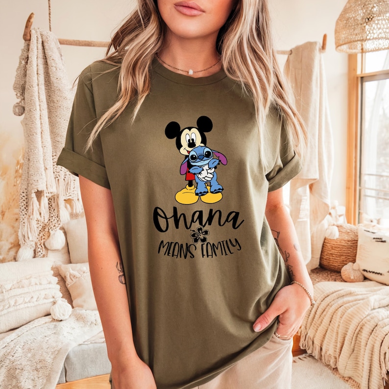 May include: Olive green t-shirt featuring a graphic of Mickey Mouse hugging Stitch. The text "Ohana Means Family" is below the characters. The shirt has rolled-up sleeves.