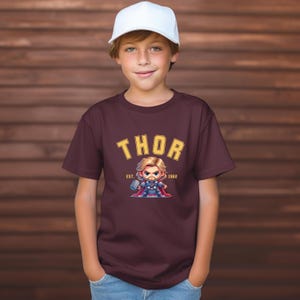 K&ouml;nnte beinhalten: Ein weinrotes T-Shirt mit dem Wort "THOR" in gelben Blockbuchstaben und einer Cartoon-Illustration von Thor. Die Illustration zeigt Thor mit einem Hammer und einem Umhang. Der Text "EST. 1962" steht darunter. Die Person tr&auml;gt eine wei&szlig;e Baseballkappe.